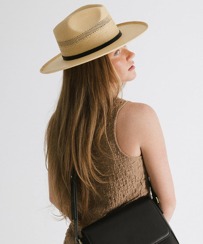 Woman with long hair wearing a natural colored straw fedora hat with a hat band and a brown top, carrying a black bag against a plain background #color_natural