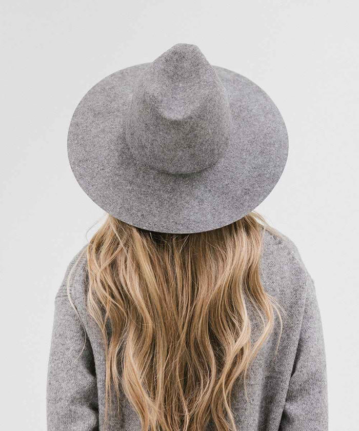 Woman with long blonde hair wearing a grey wide brim fedora hat and a grey sweater, facing behind against a plain background #color_grey