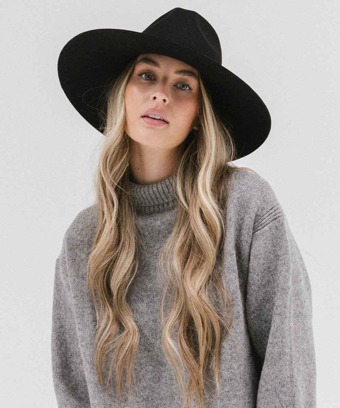 Woman wearing a black wide brim fedora hat and a grey sweater against a plain background #color_black
