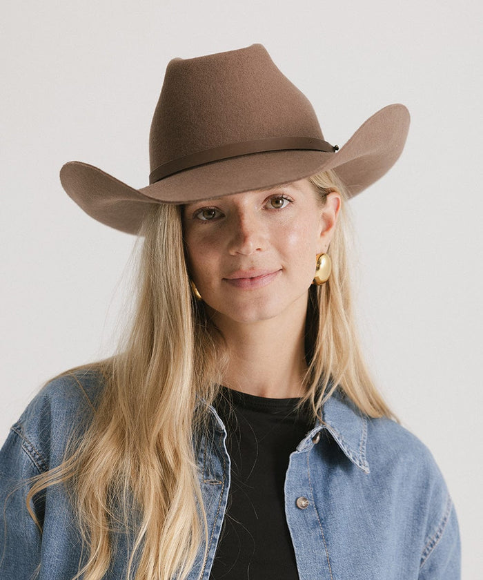 Woman wearing a chocolate colored cowboy hat with an upturned brim and an attached leather band, and a black t-shirt with a denim shirt against a white background #color_chocolate