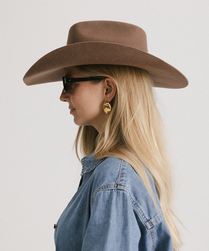 Woman wearing a chocolate colored cowboy hat with an upturned brim and an attached leather band, sunglasses, and a denim shirt, facing sideways against a white background #color_chocolate