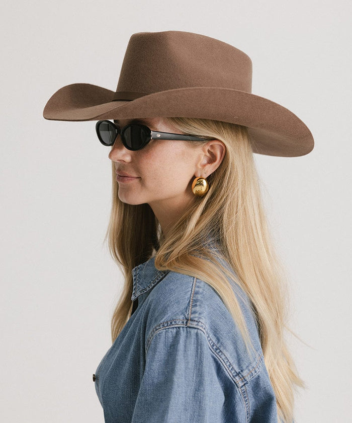 Woman wearing a chocolate colored cowboy hat with an upturned brim and an attached leather band, sunglasses and a denim shirt, looking sideways against a white background #color_chocolate
