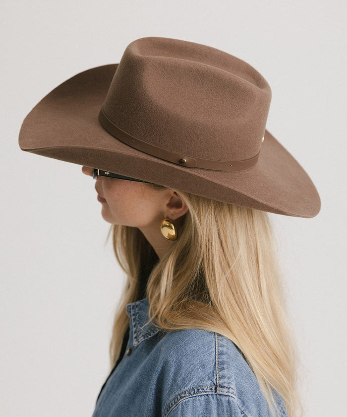 Woman wearing a chocolate colored cowboy hat with an upturned brim and an attached leather band, and a denim shirt, facing sideways against a white background #color_chocolate