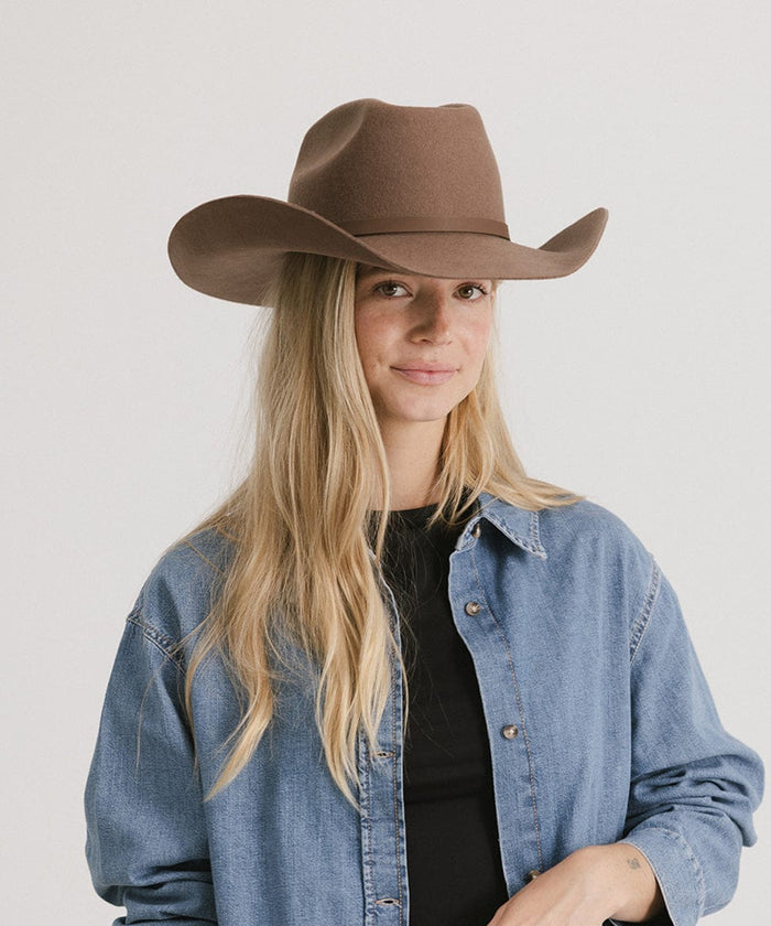Woman wearing a chocolate colored cowboy hat with an upturned brim and an attached leather band, and a black t-shirt with a denim shirt against a white background #color_chocolate