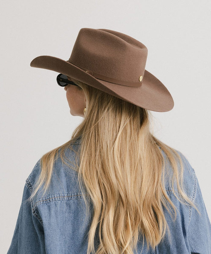 Woman wearing a chocolate colored cowboy hat with an upturned brim and an attached leather band, and a denim shirt, facing behind against a white background #color_chocolate