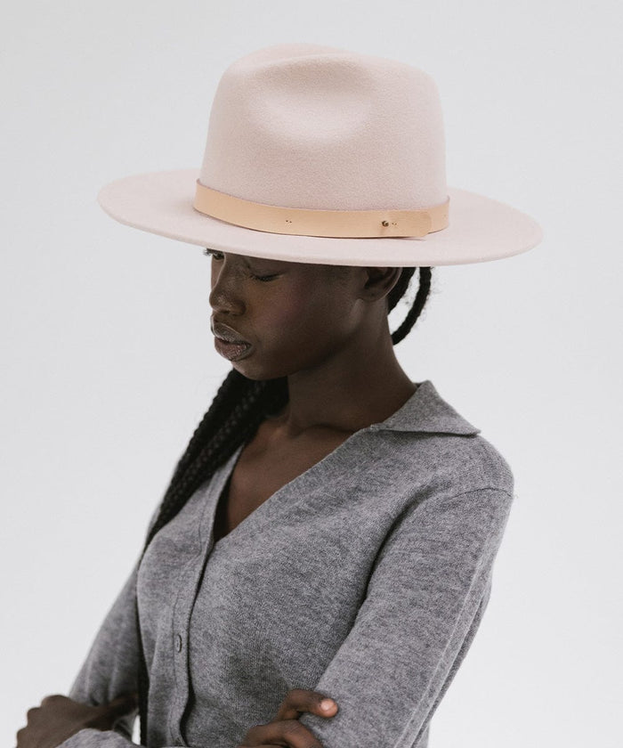 Woman wearing a blush pink colored fedora hat with a hat band and a grey shirt, looking sideways against a plain background  #color_blush pink