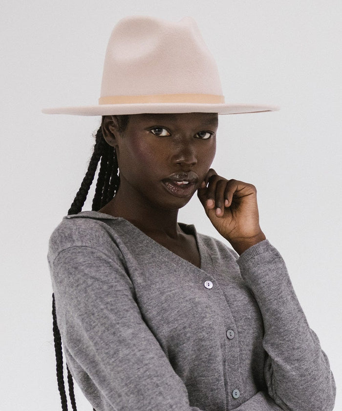 Woman wearing a blush pink colored fedora hat with a hat band and a grey shirt against a plain background  #color_blush pink