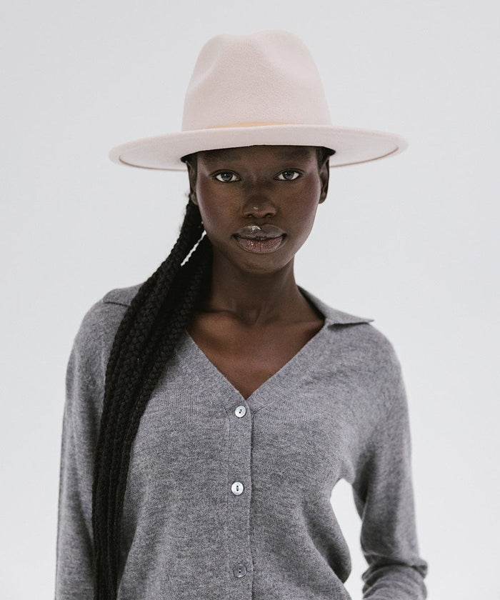 Woman wearing a blush pink colored fedora hat with a hat band and a grey shirt against a plain background  #color_blush pink