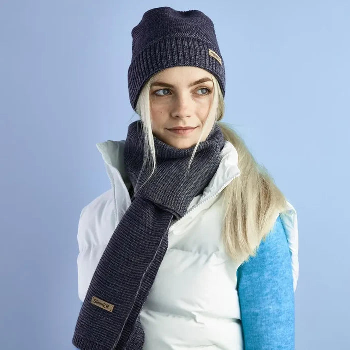 Sinner Brighton wide and long acrylic scarf in blue being worn by a woman waering a body warmer and a beanie hat