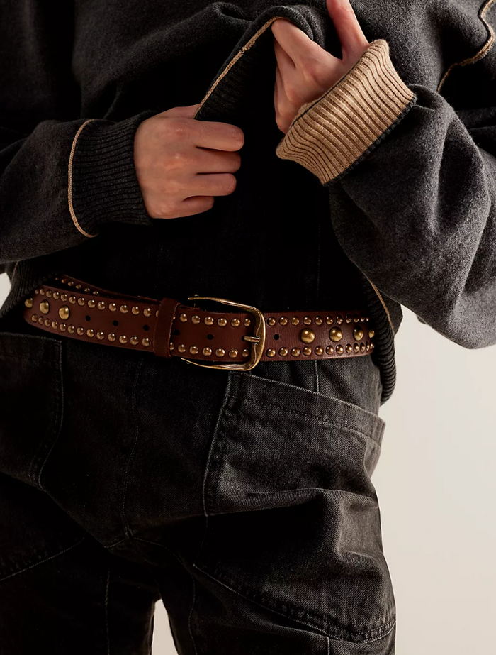Person wearing a dark jacket with a studded belt and beige cuffs on a light background