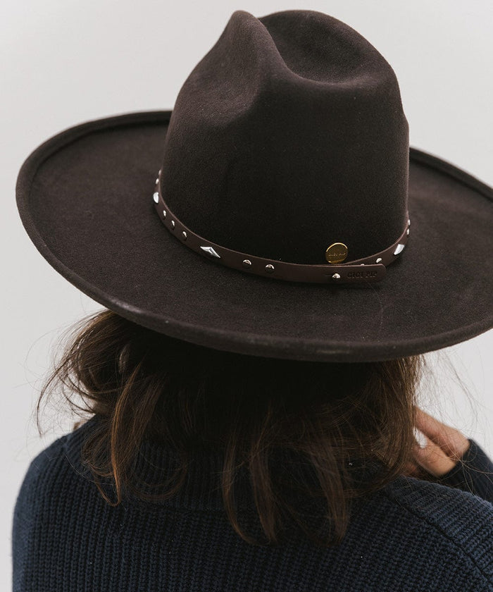 Woman wearing a brown hat with a chocolate colored studded leather hat band against a plain background #color_chocolate