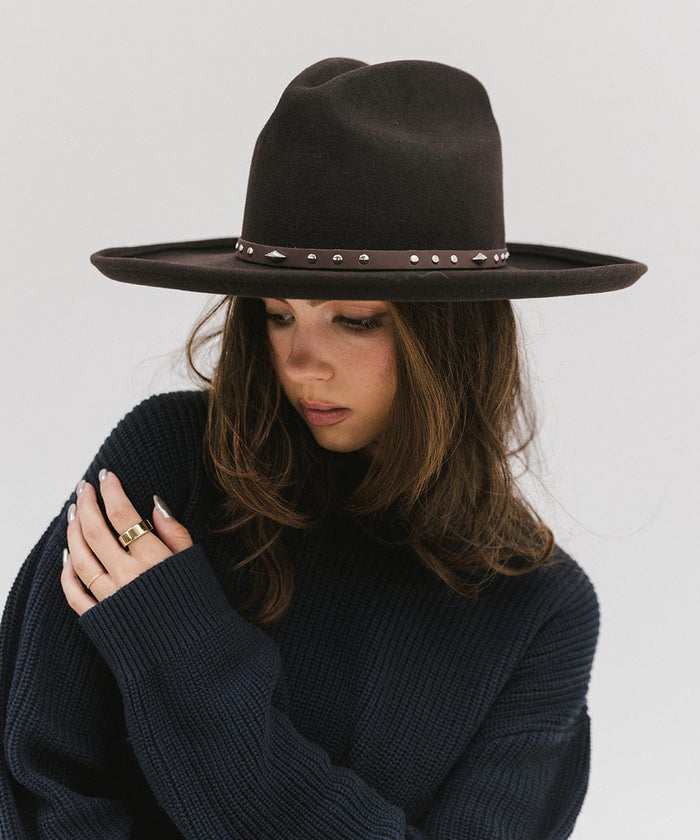 Woman wearing a brown hat with a chocolate colored studded leather hat band and a blue sweater against a plain background #color_chocolate