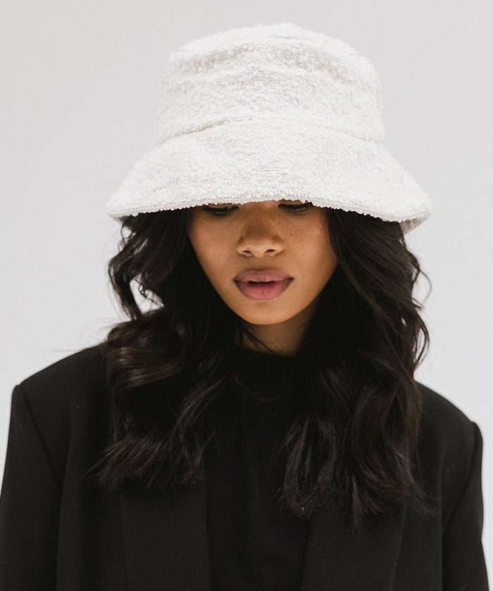 Woman wearing a white bucket hat and a black t-shirt with a black blazer, looking down against a plain background  #color_white