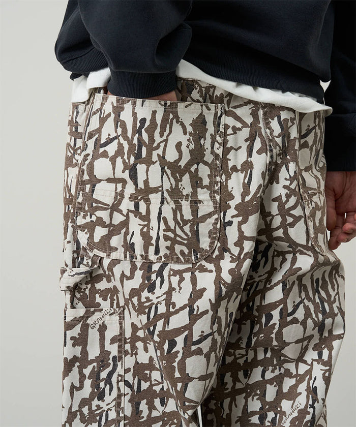 Back view of a model wearing grey reed camo Gramicci Taos Canvas Pant and a blue jacket on a light-tone background.
