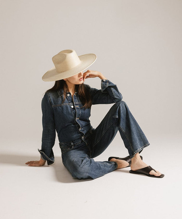 Woman wearing a natural colored western wide brim straw hat, a denim jacket, jeans and black slippers, sitting against a plain background #color_natural