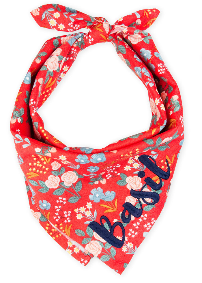A cheerful red dog bandana adorned with an all-over floral pattern of soft blooms, and leaves, tied at the top in a classic triangle shape for a fun, fashionable flair. The name "Basil" is embroidered on the bottom right.