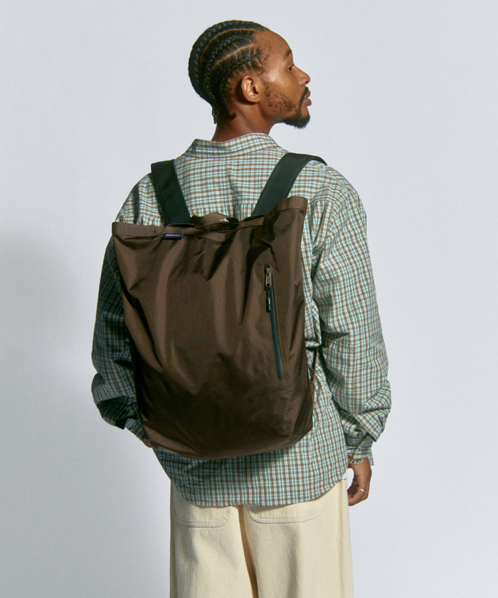 Model wearing brown Gamicci Tote Pack and a green plaid shirt on a light-tone background.