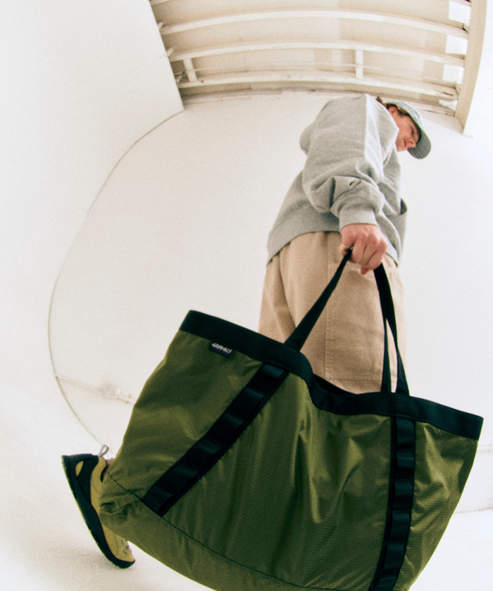 Model holding an olive green Gamicci Trail Tote and wearing a grey sweatshirt and khaki pants on a light-tone background.