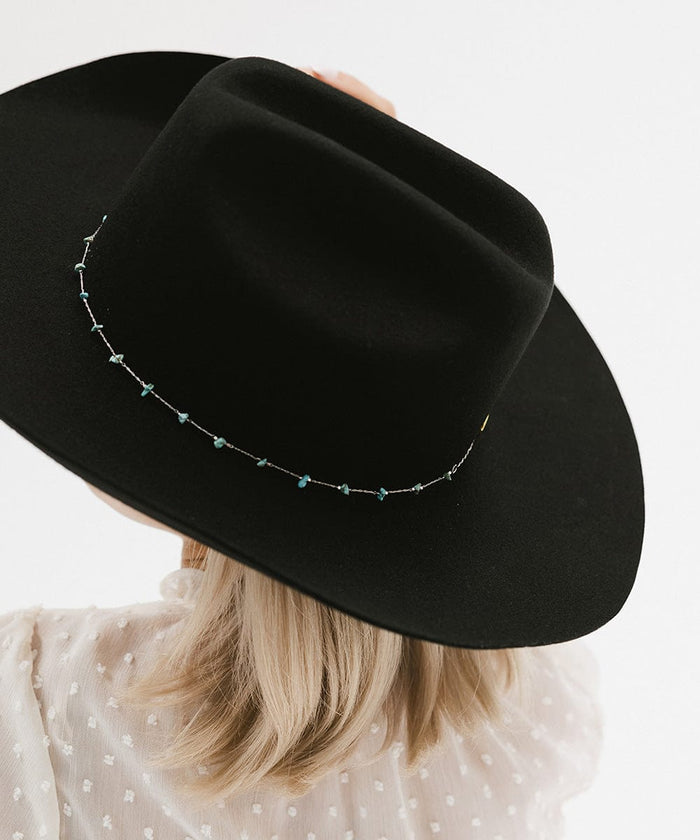 A person wearing a black hat with a turquoise stone hat band around the base and a white top, facing behind  against a plain background #color_silver