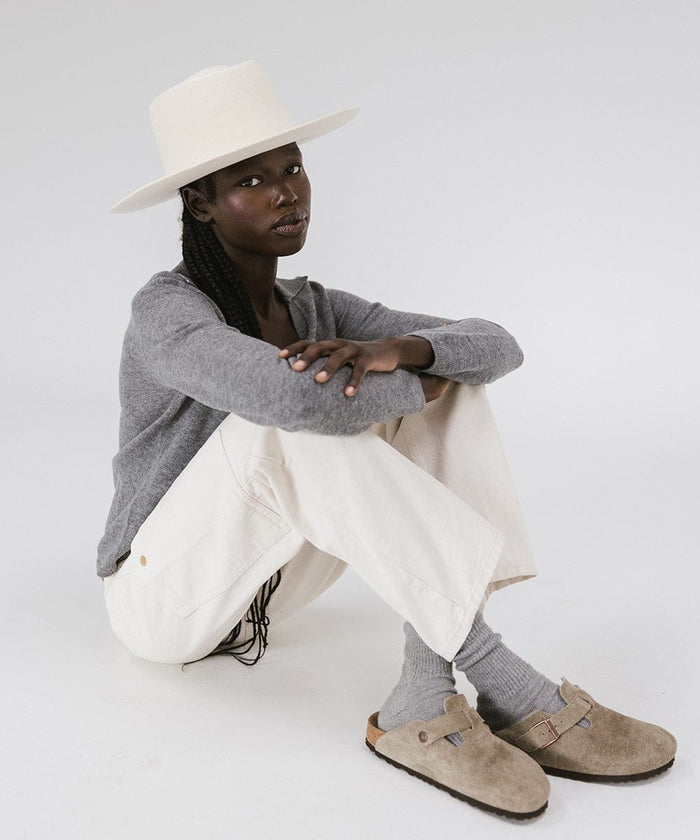 Woman wearing an off white gambler hat, a grey shirt, white pants and beige sandals, sitting against a plain background #color_off white