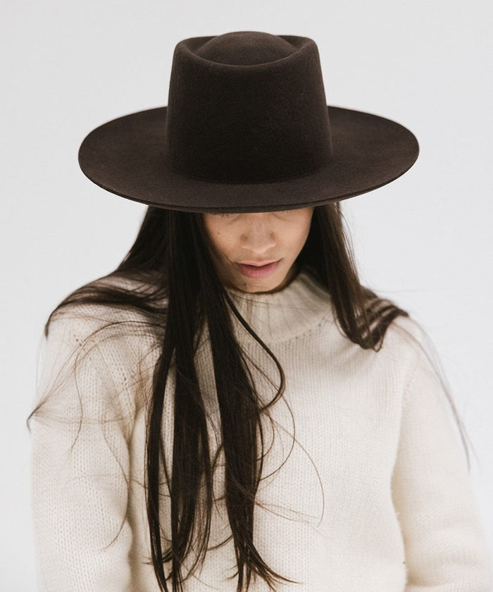 Woman wearing a dark brown gambler hat and a white sweater, looking down against a plain background #color_dark brown