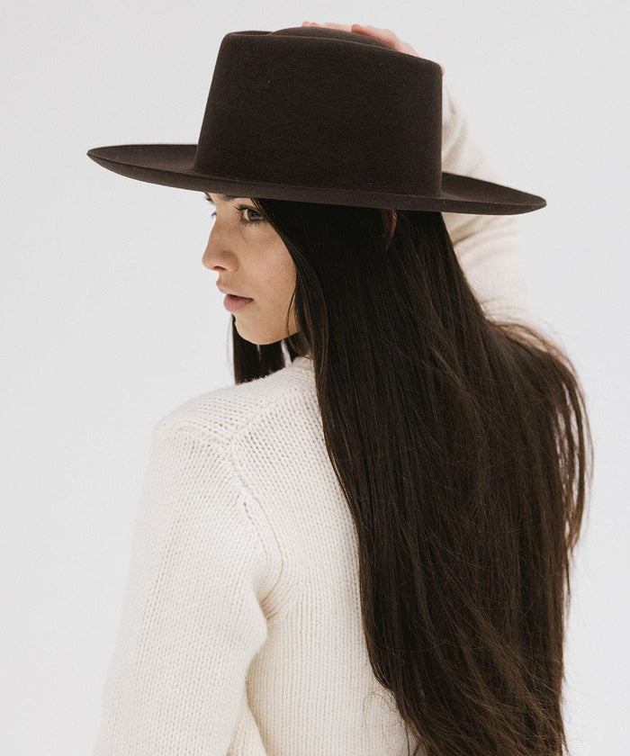 Woman with long hair wearing a dark brown gambler hat and a white sweater, looking sideways against a plain background #color_dark brown