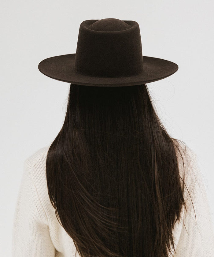 Woman with long hair wearing a dark brown gambler hat and a white sweater, facing behind against a plain background #color_dark brown