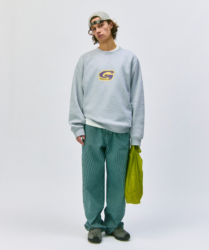 Model wearing a grey sweatshirt and green pants holding an olive Gramicci Ultra-Light Shopper on a light-tone background.