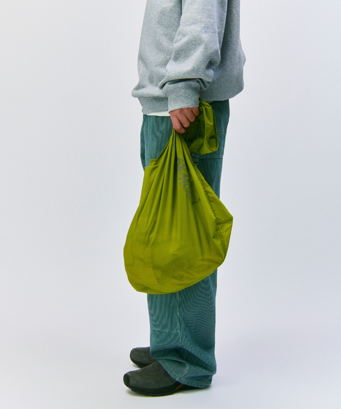 Model wearing a grey sweatshirt and green pants holding an olive Gramicci Ultra-Light Shopper on a light-tone background.