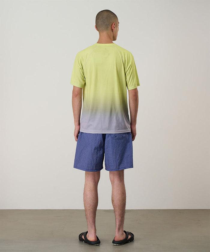 Back view of a model wearing a yellow gradient Gramicci UPF Shield S/S Top with a white Gramicci graphic and blue shorts on a light-tone background.