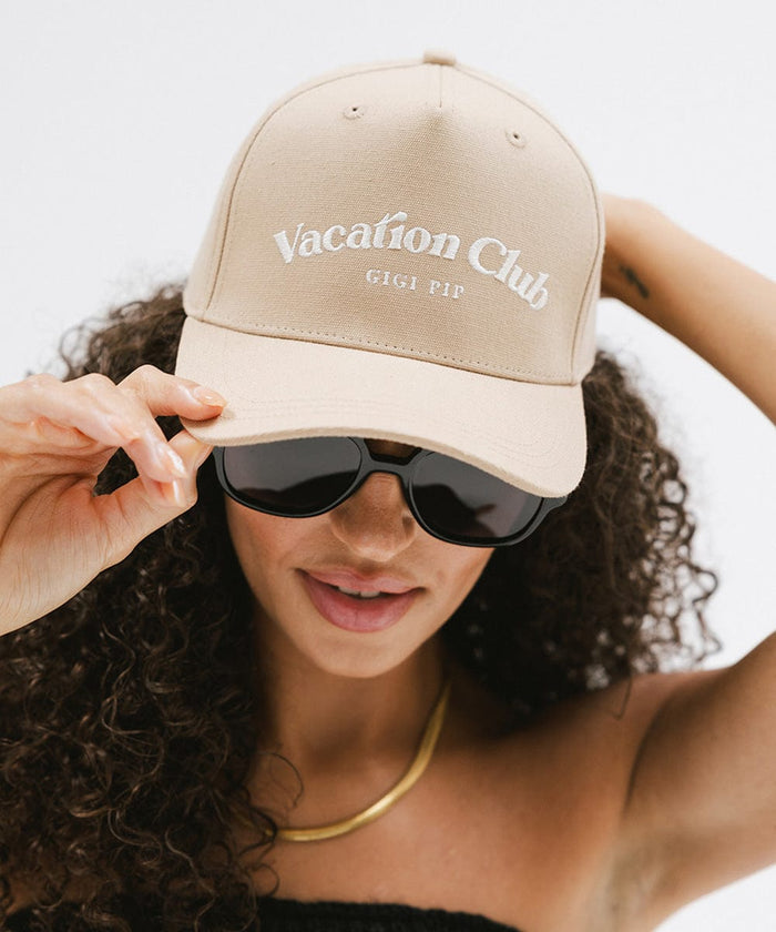 Woman wearing a tan cap with 'Vacation Club' text and sunglasses against a plain background  #color_tan