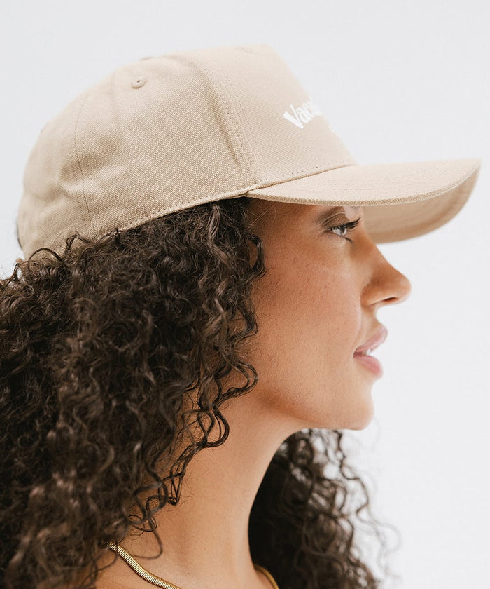 Woman wearing a tan cap with 'Vacation Club' text, facing sideways  against a plain background  #color_tan