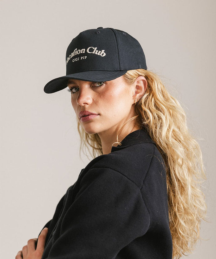 Woman wearing a navy cap with 'Vacation Club' text and a black shirt, posing sideways against a plain background  #color_navy