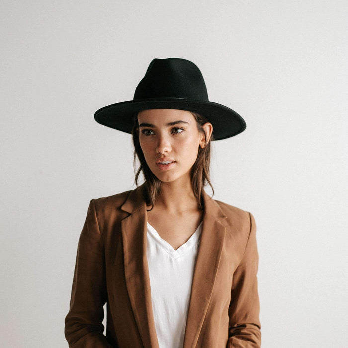 Woman wearing a black fedora hat and a white t-shirt with a brown coat against a plain background #color_black