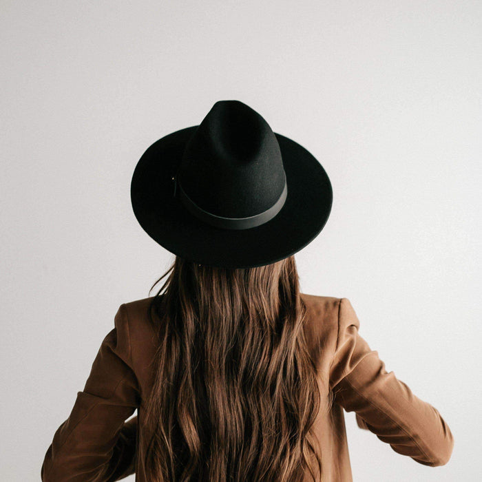 Woman wearing a black fedora hat with a hat band and a brown coat, facing behind against a plain background #color_black