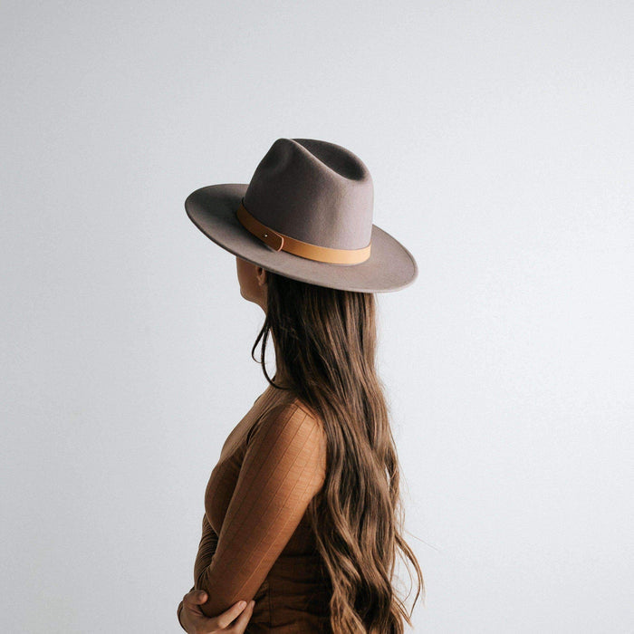 Woman wearing a light grey fedora hat with a hat band and a brown top, posing sideways against a plain background #color_light grey