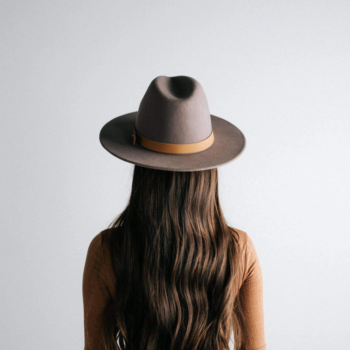 Woman wearing a light grey fedora hat with a hat band and a brown top, facing behind against a plain background #color_light grey