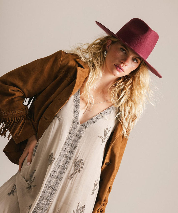 Woman wearing a brown jacket, white dress with black patterns, and a merlot fedora hat against a plain background #color_merlot