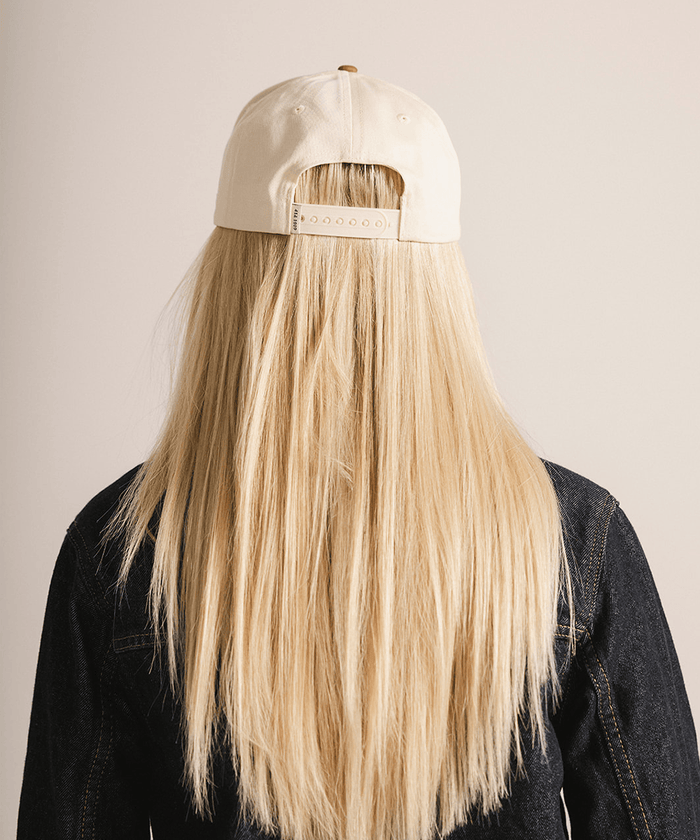 Person wearing a beige cap with a brand logo, obscuring their face, on a plain background. #color_cream - caramel
