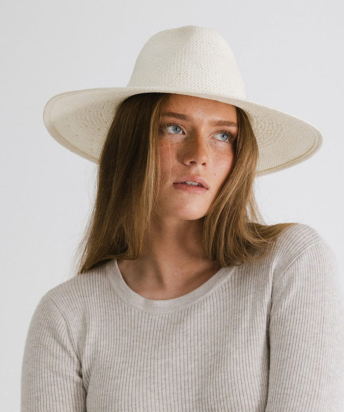 Woman wearing a white wide brim fedora straw hat and a grey top, looking sideways against a plain background  #color_white