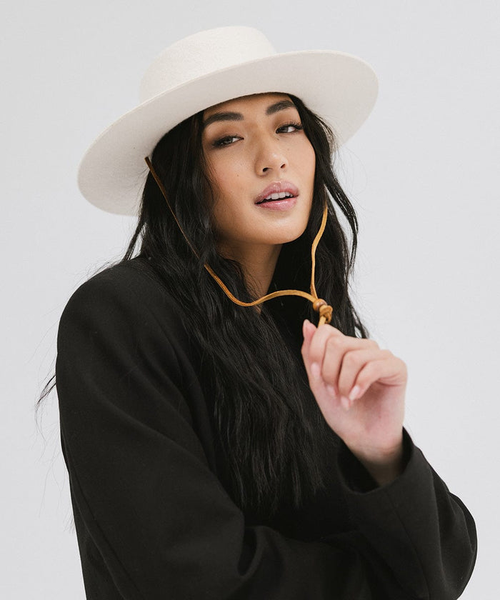 Woman wearing an off white wide-brimmed hat with a strap and a black coat against a plain background #color_off white