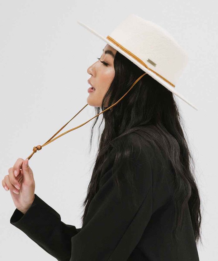 Woman wearing an off white wide-brimmed hat with a strap and a black coat, facing sideways against a plain background #color_off white