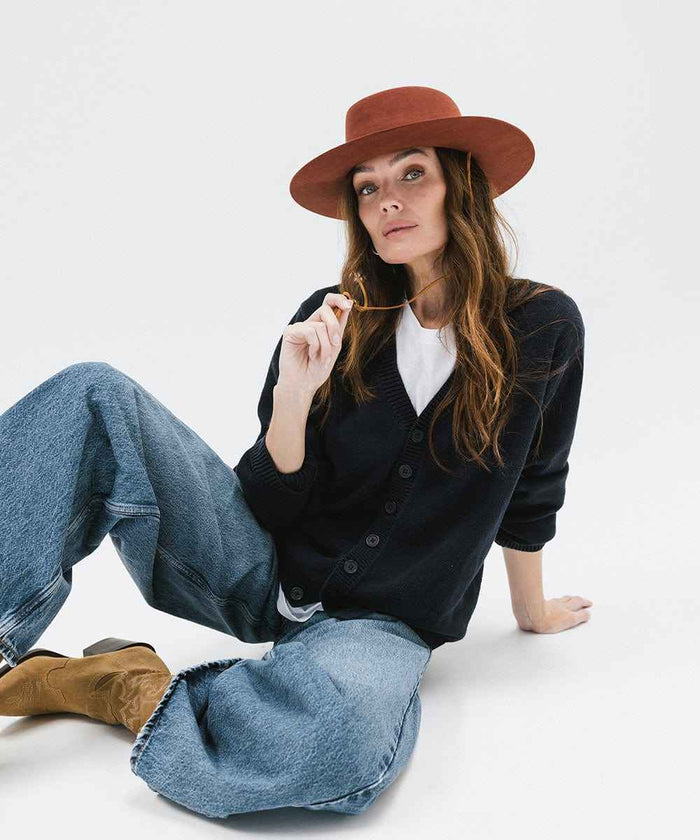 Woman wearing a dark blue shirt, blue jeans, and a rusty red colored hat, sitting against a white background #color_rusty red