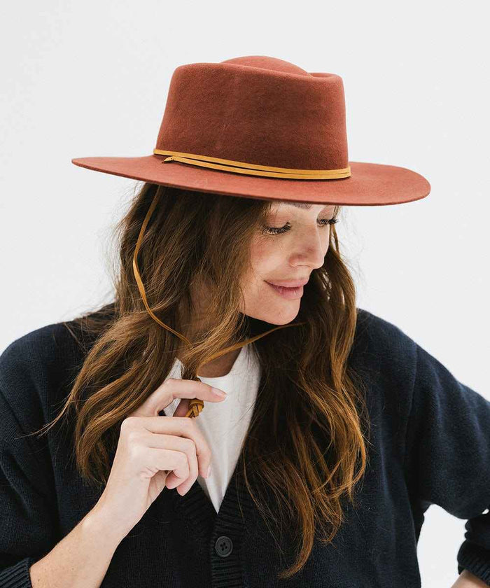 Woman wearing a rusty red colored hat with a yellow band and a dark blue shirt, looking sideways against a white background #color_rusty red