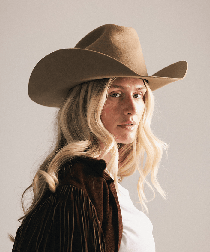 A woman with blonde hair wearing a brown cowboy hat and a brown fringed jacket over a white shirt, photographed against a plain light gray background #color_brown