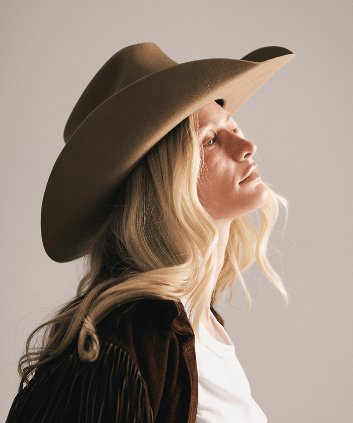 A woman in profile wearing a wide-brimmed brown cowboy hat, a brown fringed jacket, and a white top against a plain light gray studio background #color_brown