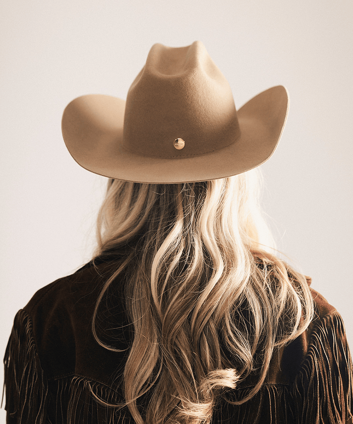 A woman viewed from behind wearing a tan felt cowboy hat with a small gold button and a brown fringed jacket, against a plain light beige background #color_brown