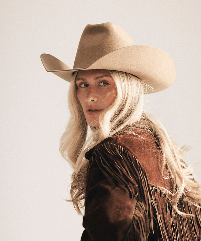 A woman with long blonde hair wearing a tan cowboy hat and a brown fringed suede jacket, photographed against a plain light gray studio background #color_brown