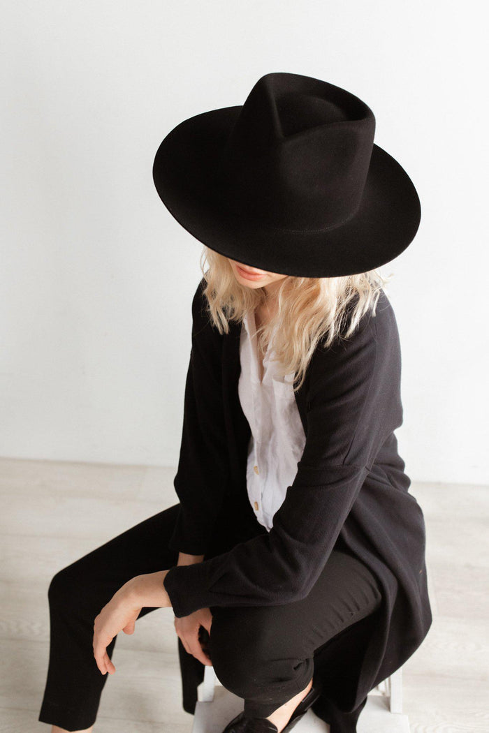 Woman wearing a black fedora hat, a white shirt with a black coat and black pants, sitting against a plain background #color_black