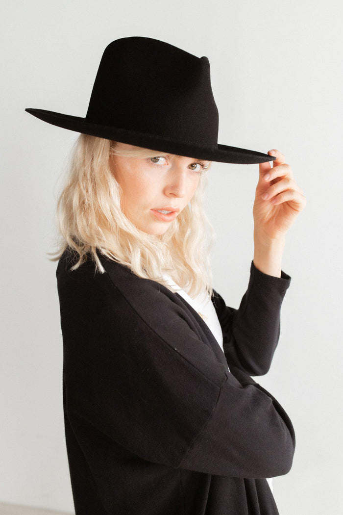 Woman wearing a black fedora hat and a white shirt with a black coat against a plain background #color_black